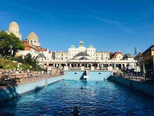 Soligt utomhuspool vid ett termalbad i Budapest. Människor solar och badar i det blå vattnet framför en imponerande byggnad.