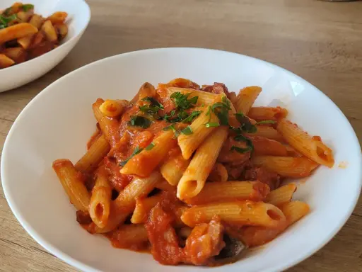 Penne z sosem pomidorowym i kawałkami kiełbasy, posypane natką pietruszki. Pyszny makaron z kiełbasą na białym talerzu.