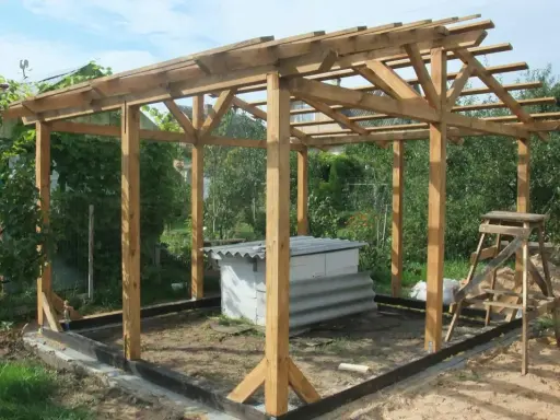 Drewniana altanka z palet w budowie, z dachem i słupami. W środku betonowa konstrukcja.