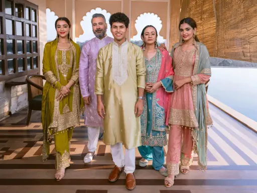 A family poses in vibrant traditional clothing from India. The women wear ornate salwar kameez sets in olive green, teal, and pink, while the men are dressed in elegant kurtas.
