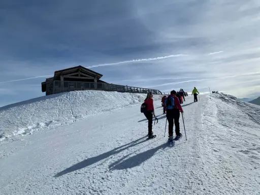 Gruppo di escursionisti con ciaspole su un sentiero innevato, con un rifugio in legno in cima a una collina. Posti belli in montagna Piemonte inverno.