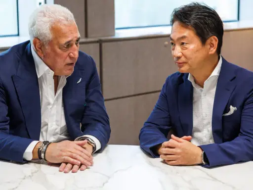 Koji Watanabe conversa con un hombre mayor, ambos con trajes azules. Un modelo de coche deportivo negro se ve en el fondo.