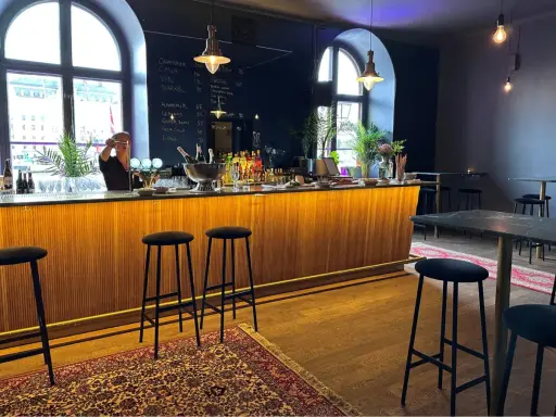En bar med träpanel och svarta barstolar. En person bakom baren förbereder en drink. En meny på svarta tavlan visar priser för drycker i det musikaliska kvarteret.