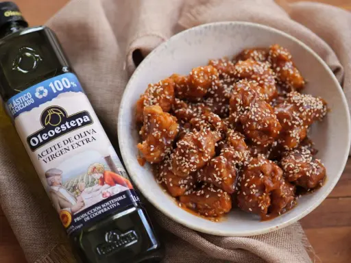 Delicioso pollo agridulce cubierto de semillas de sésamo en un cuenco, junto a una botella de aceite de oliva virgen extra Oleoestepa.
