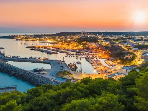 Porto illuminato al tramonto, un invito al turismo Salento. Barche ormeggiate e case che brillano sulla costa.