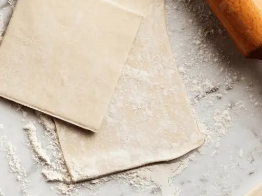 Pâte feuilletée prête à être travaillée, saupoudrée de farine sur un plan de travail en marbre. Un rouleau à pâtisserie en bois est visible sur la droite.