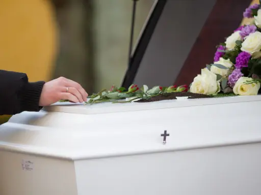 Dłoń z obrączką kładzie róże na białej trumnie, ozdobionej bukietem kwiatów i krzyżem.
