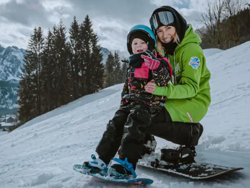 Uśmiechnięta instruktorka na stoku narciarskim dla dzieci trzyma małego snowboardzistę.