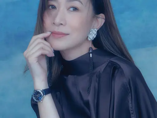 A woman with a thoughtful expression, her finger near her lips, wears a dark satin dress, a striking earring, and a blue-faced watch. She looks xa thi man.