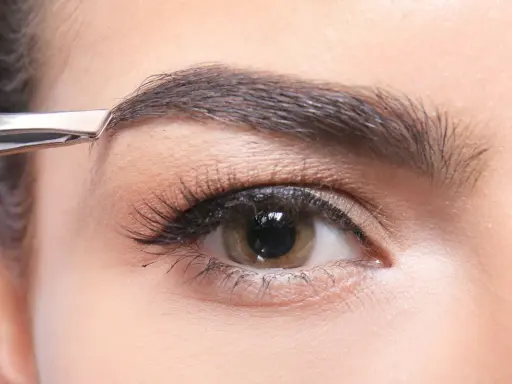 Une pince à épiler retire délicatement un poil du sourcil, affinant la ligne pour un regard parfait.