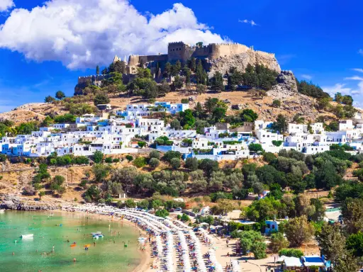 En vitkalkad by klättrar uppför en kulle mot en borgruin. Nedanför breder en strand ut sig med solstolar och turkost vatten. En vy över Rhodos karta Europa.