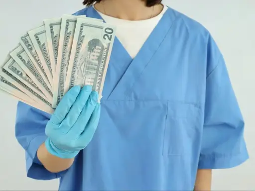 Higienistka stomatologiczna w niebieskim fartuchu pokazuje wachlarz banknotów 20 dolarowych. Zarobki netto mogą być satysfakcjonujące.