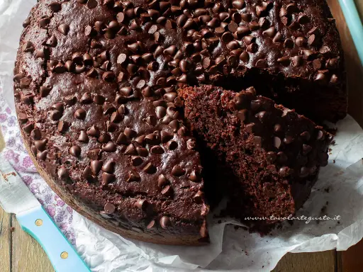 Torta al cioccolato senza uova, soffice e golosa, ricoperta di gocce di cioccolato. Una fetta mostra la sua morbidezza.