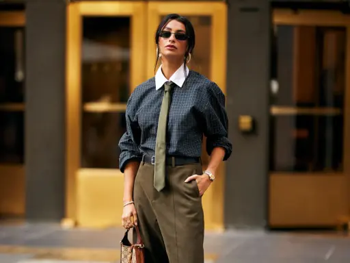 Donna con un audace outfit con cravatta, camicia a quadri, pantaloni kaki e borsa di lusso.