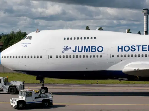 En person sitter på taket av ett jumbo stay. Flygplanet är vitt med blå detaljer och texten "JUMBO HOSTEL" syns tydligt.