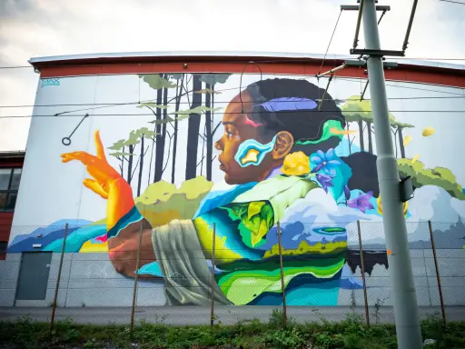 Färgstark street art på en vägg visar en person i profil med en fjäder i håret, omgiven av träd och blommor.