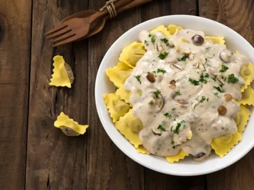 Ravioli w kremowym sosie śmietanowym z grzybami i natką pietruszki, podane na drewnianym stole.