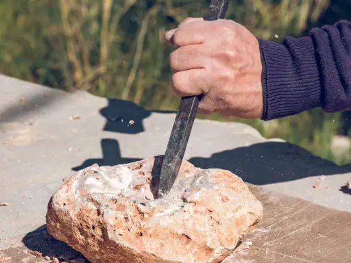 Dłoń z dłutem pracuje nad kamieniem, tworząc pył. To jak wiercenie w granicie z udarem czy bez – wymaga precyzji i siły.
