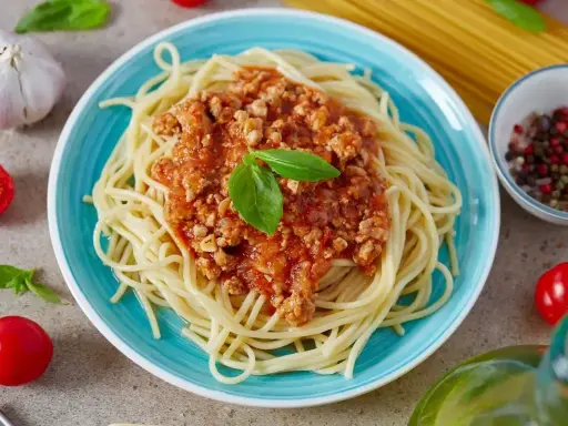 Pyszne spaghetti z bogatym sosem bolognese, udekorowane listkiem bazylii. Obok czosnek, pomidorki i przyprawy.