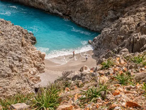 Malownicza zatoka z turkusową wodą i piaszczystą plażą, otoczona skalistymi klifami. Dwie osoby spacerują po brzegu. Chania plaże zachwycają.