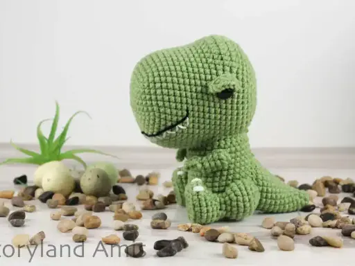 A green crocheted dinosaur sits on a white surface surrounded by small stones and a small plant.