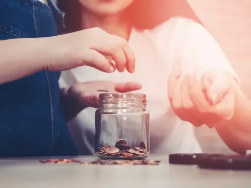 Dwie osoby wrzucają monety do słoika, oszczędzając na przyszłość. Zastanawiasz się, ile wynoszą alimenty na studenta?