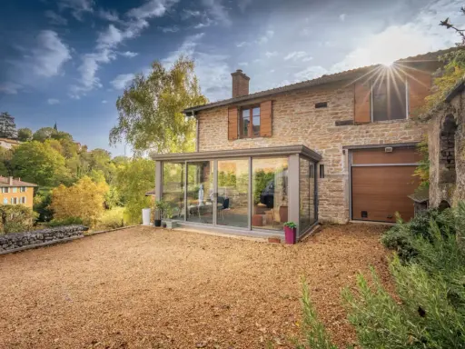 Maison en pierre avec véranda moderne, illustrant la différence entre véranda et extension. Le soleil brille sur le paysage verdoyant.
