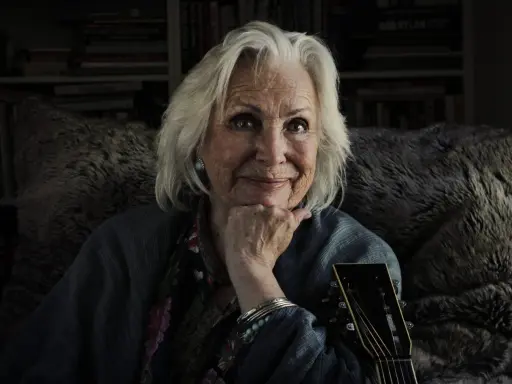 Äldre kvinna med grått hår, gitarr och en fluffig pläd. Trots att Marie Bergman är sjuk, lyser hennes ögon av liv.
