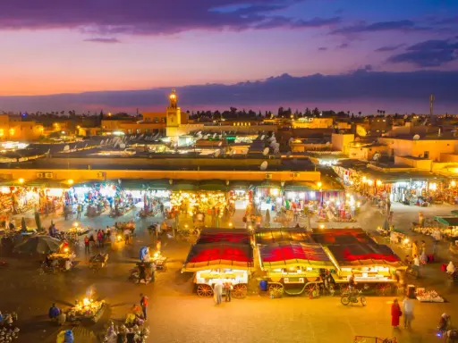 Der belebte marrakesch marktplatz erwacht bei Dämmerung zum Leben. Stände mit bunten Waren, Essensverkäufer und Menschenmassen beleben die Szene unter dem lila Himmel.