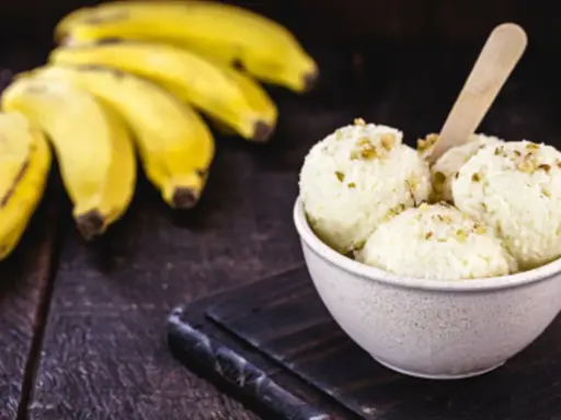 Delicioso helado de plátano cremoso, adornado con nueces, junto a racimos de plátanos frescos sobre una mesa de madera rústica.