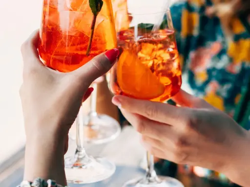 Zwei Hände halten Aperol Spritz Gläser mit Eis und Orangenscheiben. Ein perfektes Aperol Rezept für den Sommer.