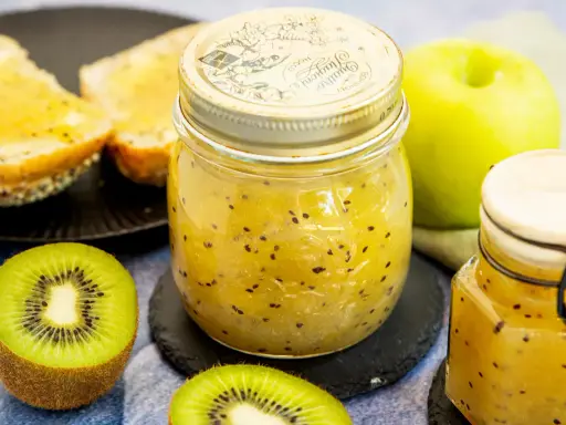 Ein Glas mit leuchtend gelber kiwi-marmelade mit schale, daneben frische Kiwischeiben und geröstetes Brot.