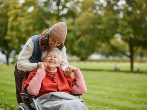Una giovane donna sorride mentre abbraccia un'anziana in sedia a rotelle in un parco. Un momento di gioia che mostra come un caregiver può sopravvivere con amore.