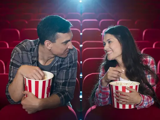 Para w kinie, zajadając popcorn, patrzy na siebie z uśmiechem. Ich spojrzenia sugerują, że to nie tylko film ich interesuje, ale też wzajemna bliskość.