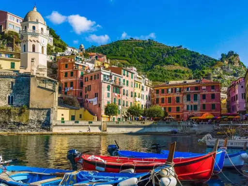 Colorate case a picco sul mare, barche rosse e blu in primo piano, un campanile svetta sulla collina. Un sogno di vacanze 5 Terre.