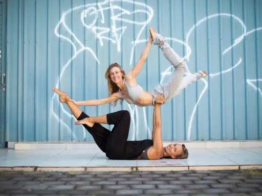 Två kvinnor utför acro yoga framför en blå vägg med graffiti. Den ena ligger på rygg och lyfter den andra som är i en böjd position.
