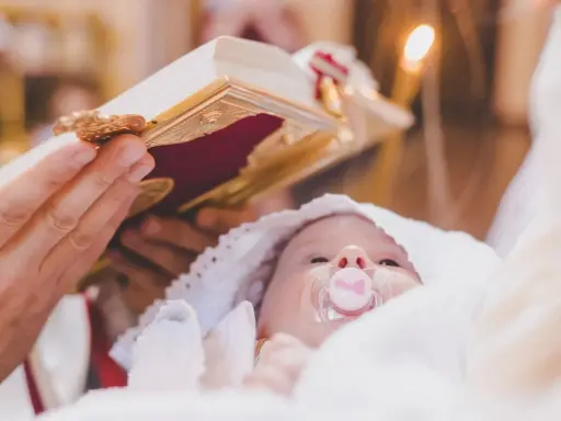 Dziecko z chupkiem w buzi, otulone białym materiałem, podczas ceremonii chrztu. Nad nim trzymana jest księga, a w tle płonie świeca.