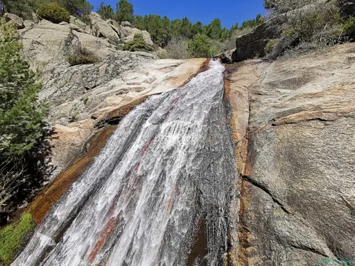 El agua desciende por la roca en la cascada del Hornillo, rodeada de vegetación y rocas graníticas bajo un cielo azul.