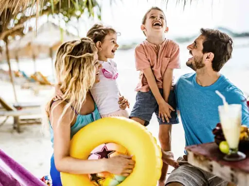 Familie lacht am Strand, genießt die Sommerferien. Ein gelber Schwimmring und Cocktails deuten auf Urlaubsfreude in den Bundesländern hin.