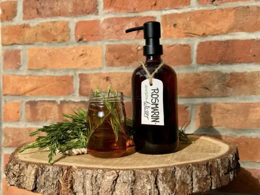 Zutaten für Rosmarinwasser Haare selber machen: frischer Rosmarin, ein Glas mit Flüssigkeit und eine dunkle Flasche mit Pumpspender.