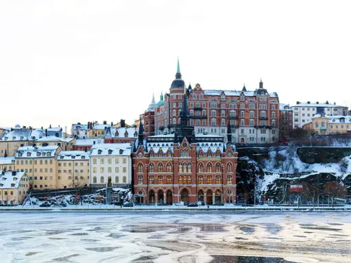 Vinter i Stockholm med snöklädda tak och isig vattenyta. Vackra byggnader reser sig mot en klar himmel.