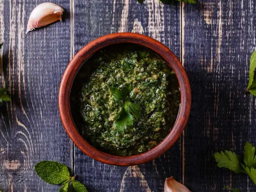 Un ciotola di terracotta contiene un pesto verde brillante, perfetto per accompagnare le acciughe al verde. Ingredienti freschi come aglio e prezzemolo circondano la ciotola.