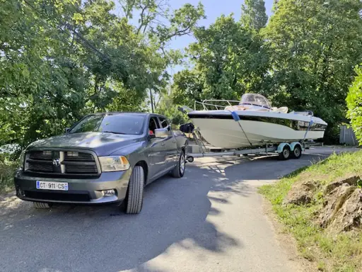 Un pick-up gris est prêt à tracter un bateau de 7m sur sa remorque, sous un ciel bleu et entouré de verdure.