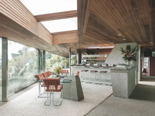 Interno della Sheats Goldstein Residence con soffitto in legno, ampie vetrate, zona pranzo con tavolo in vetro e sedie in pelle arancione, e un bancone bar in cemento con sgabelli.