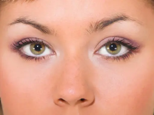 Primo piano di un viso femminile con occhi piccoli e verdi, ciglia lunghe e trucco viola sulle palpebre.