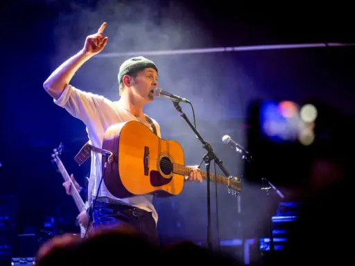 Joel Alme konsert. Sångaren med gitarr och mössa pekar uppåt under en blåbelyst spelning.
