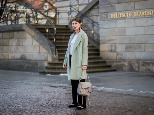 Kobieta w płaszczu w pistacjowym kolorze, z beżową torebką i ozdobnymi butami, stoi przed budynkiem Deutsche Staatsoper.