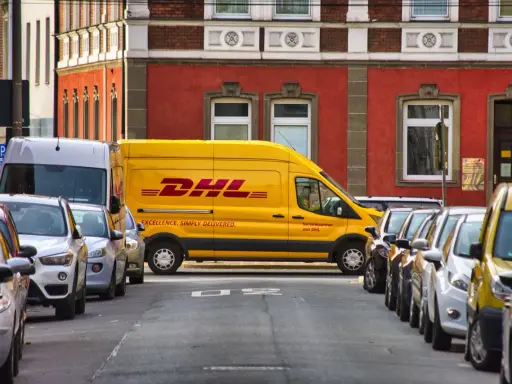 Żółta furgonetka DHL stoi zaparkowana na ulicy, obok innych samochodów. To miejsce, do którego pracują kurierzy DHL, by dostarczać paczki.