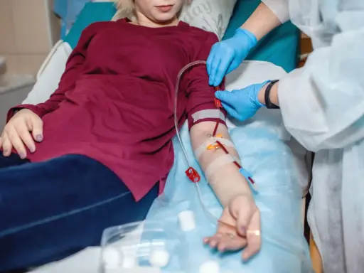 Pacjentka leży, a pielęgniarka w rękawiczkach podłącza przetokę do dializ. Krew płynie przez rurki.