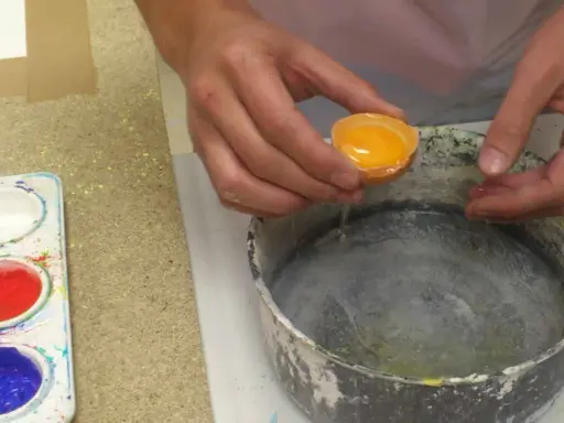 Manos vierten una yema de huevo en un recipiente para preparar una pintura al temple. Paleta con pigmentos de colores al lado.